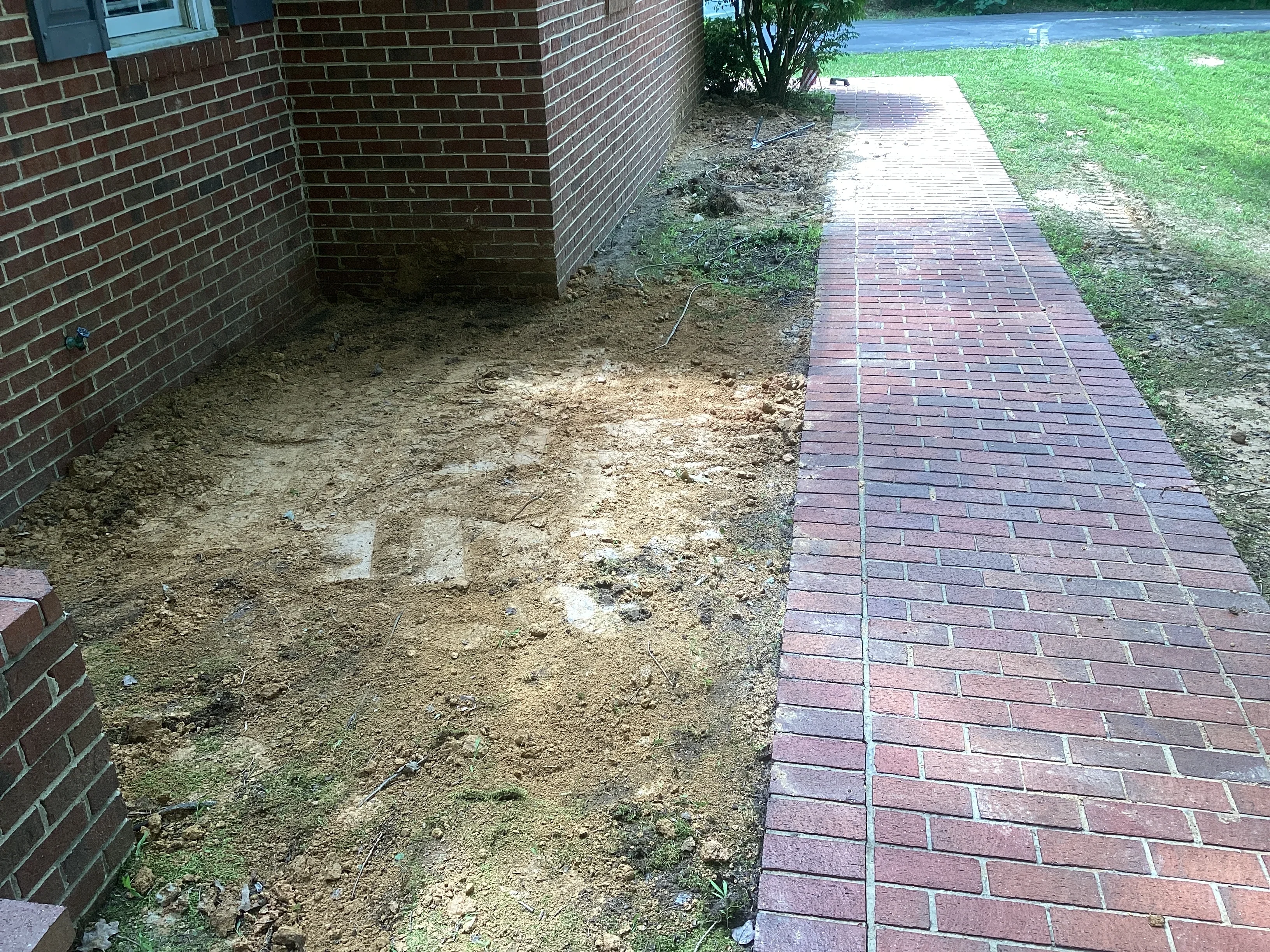 Red brick paver walkway installed next to brick home with prepared soil base visible
