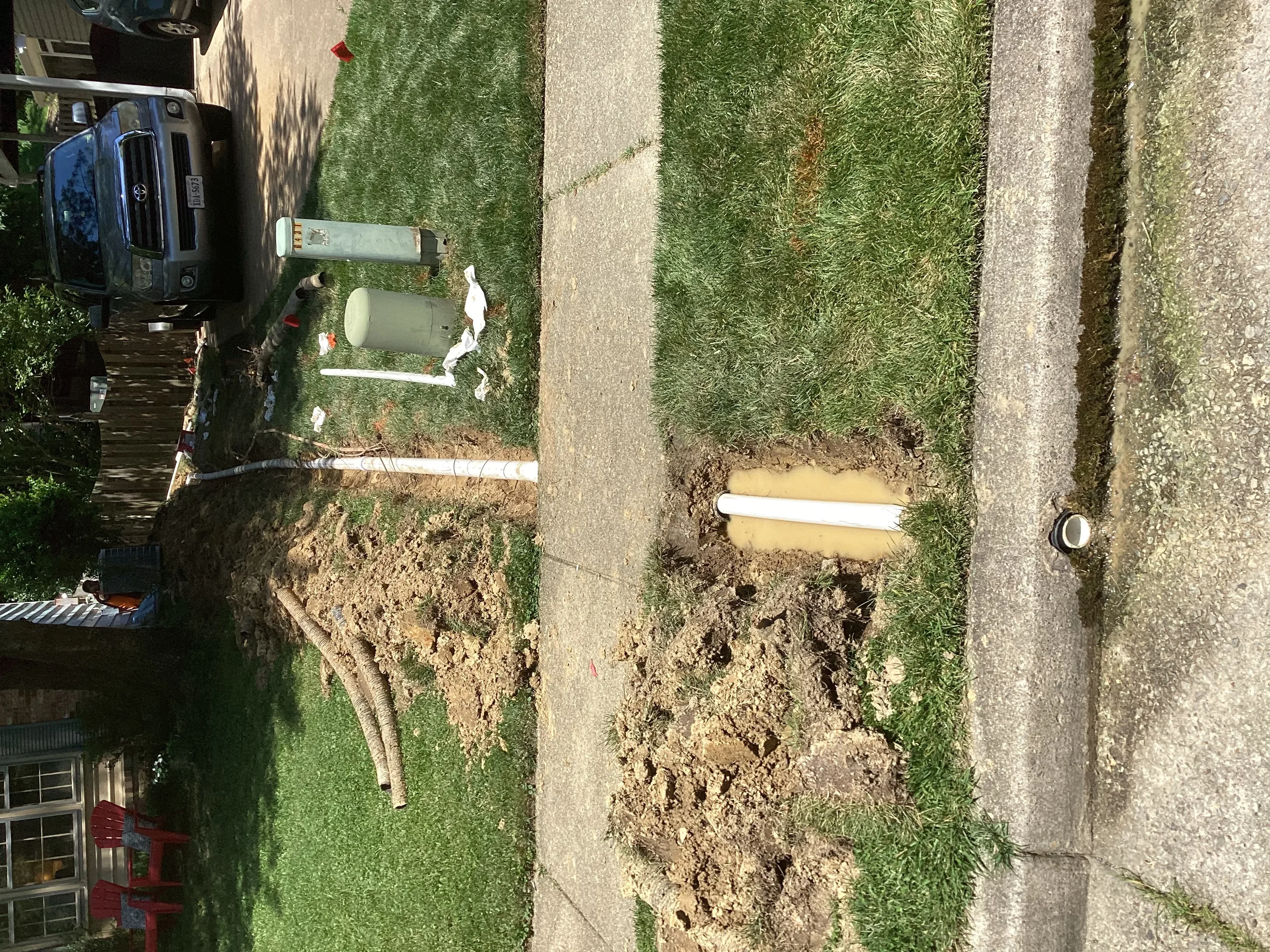 White PVC drainage pipe installed in excavated trench with soil and driveway visible