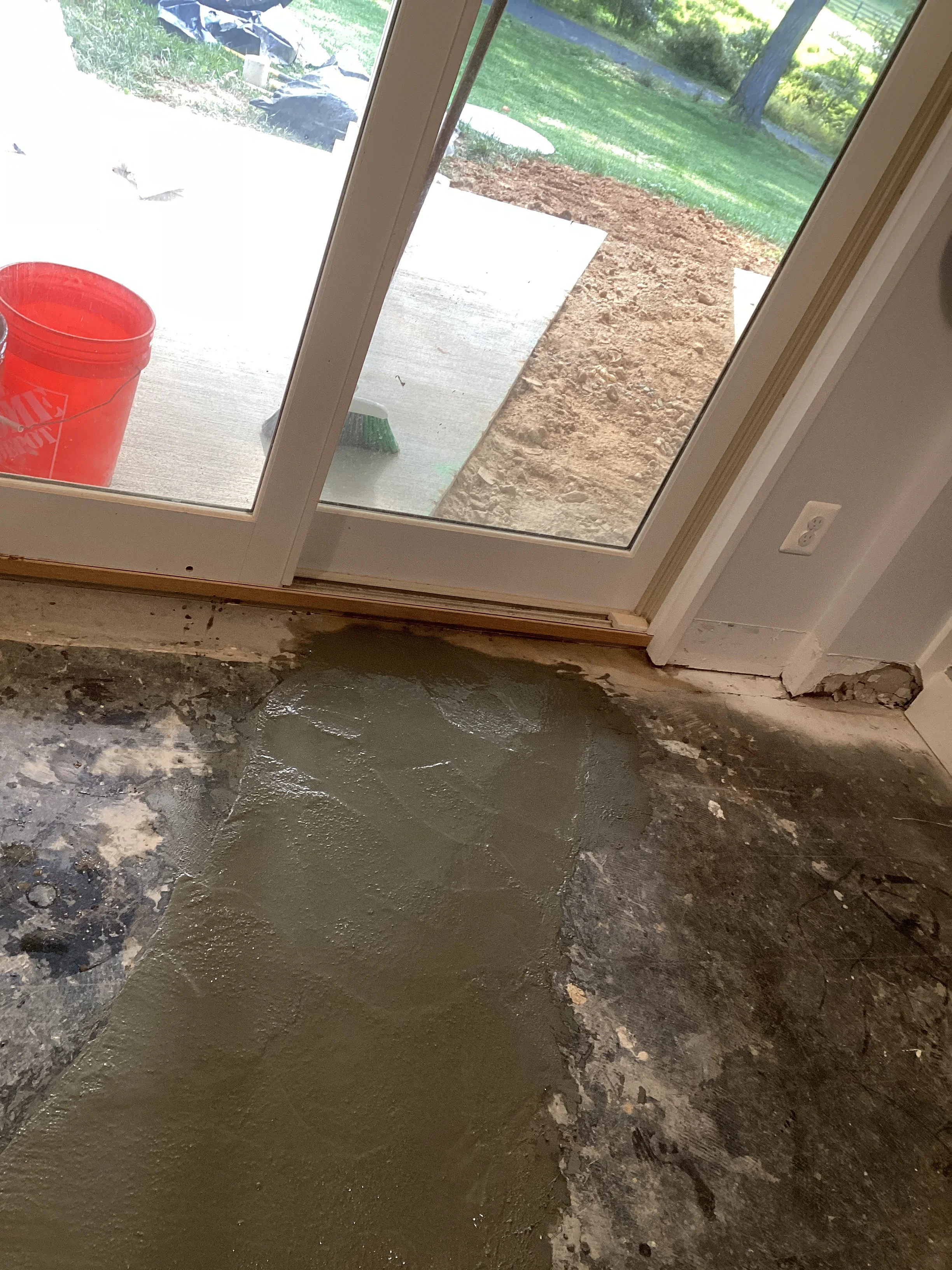 Water-damaged concrete foundation floor with wet soil visible through glass doors and exterior grading work