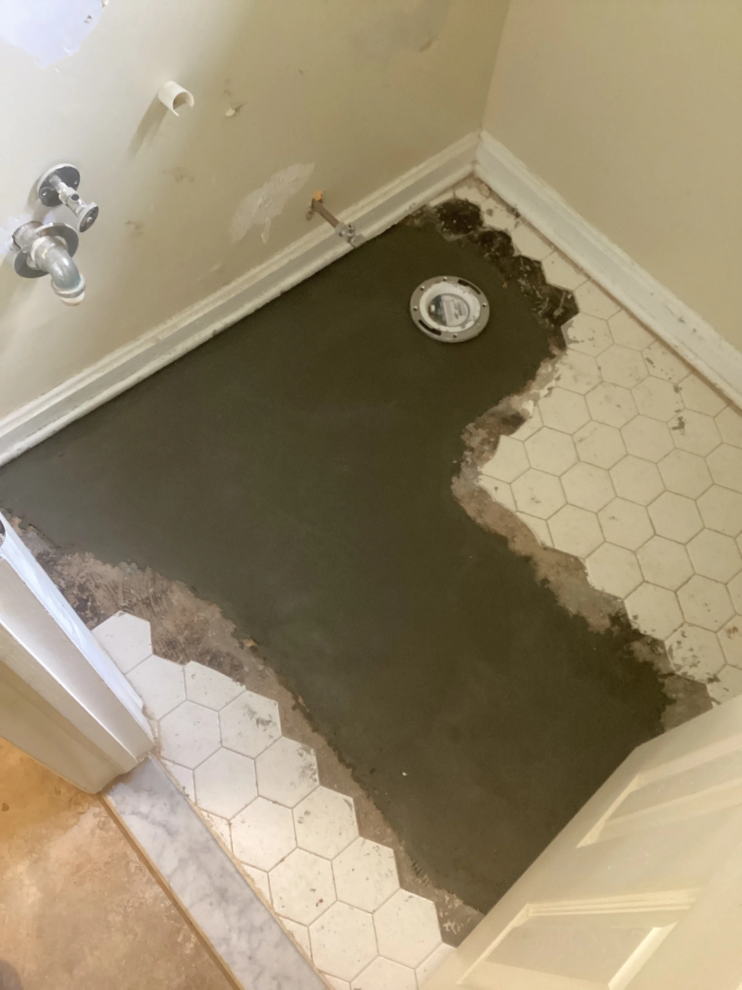 Bathroom floor with removed hexagonal tiles exposing water-damaged subfloor and plumbing access